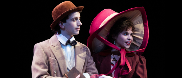 Louis Dubedat (Ian Rothauser) in a tan overcoat, black bowtie, and brown hat standing with Jennifer Dubedat (Kaitlyn Oliva) in a burgundy dress with a wide brimmed red hat tied underneath her chin for Connecticut Rep's production of A Doctor's Dilemma (Spring 2024).