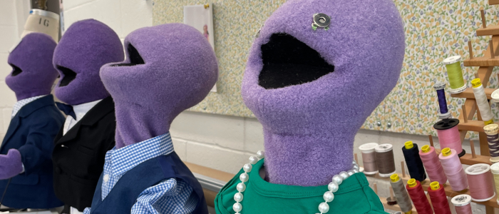 Four puppets covered in purple felt in a row in varying levels of professional attire in front of a collection of different colored threads for Connecticut Rep's War of the Worlds (Fall 2023).