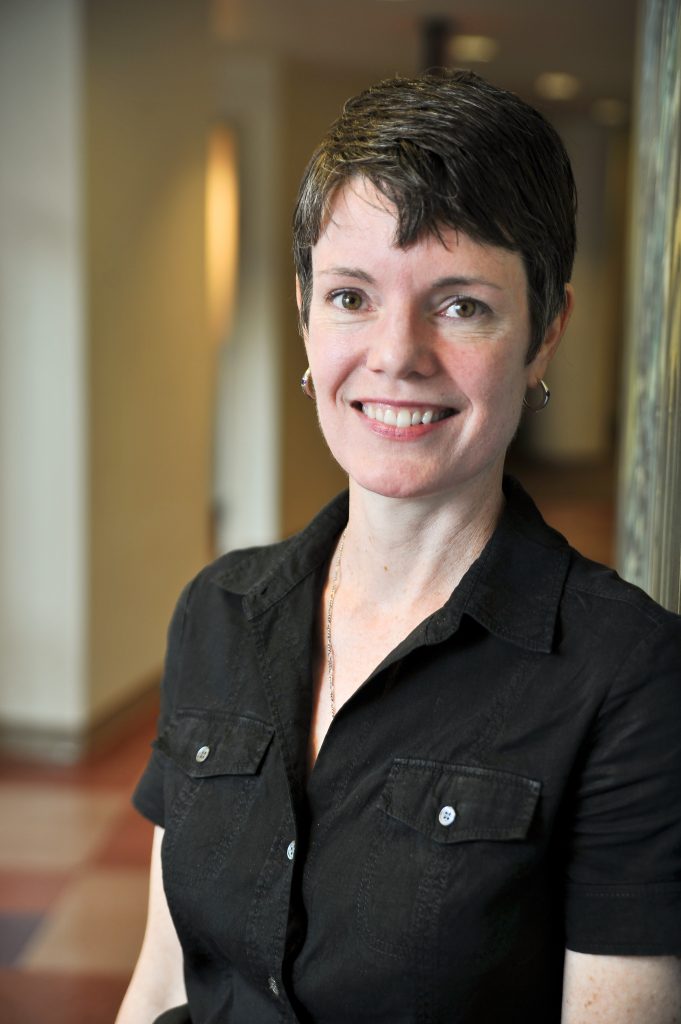 Portrait of Artistic Director Megan Monaghan Rivas- a white woman with short dark hair, wearing a black short-sleeved shirt, looking directly at the camera and smiling.