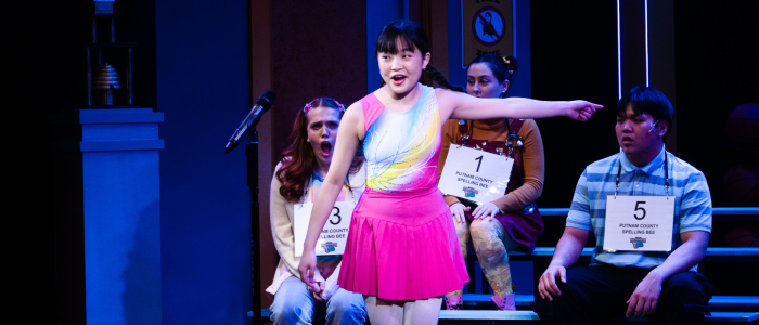 A young woman dressed in colorful gymnast outfit point to her left while youthful onlookers watch in shock. 23rd Annual Putnam County Spelling Bee.