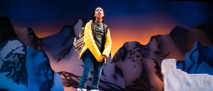 Guest Artist Téa Guarino in Connecticut Rep's A Hundred Words for Snow (Fall 2023) standing in a yellow puffer jacket in front of snow-capped mountains.