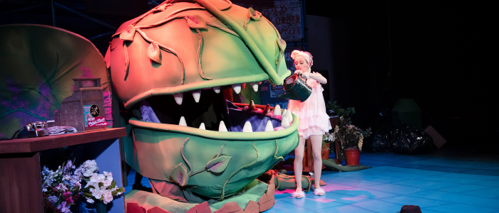 A woman in a white negligee and hair in curlers waters a very large puppet plant, in Little Shop of Horrors.