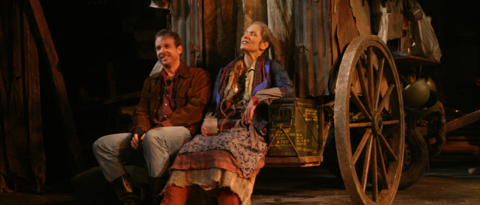 Connecticut Repertory Theatre's Mother Courage and Her Children (2004) with two actors in brown and green disheveled clothing sitting and smiling on a wagon, filled with all of their belongings.