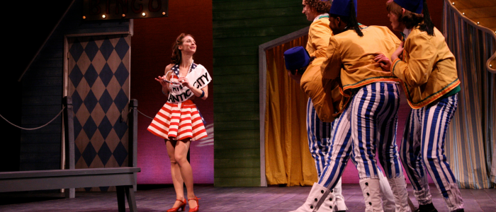 Dressed in a short red, white and blue dress and wearing a sash reading "Miss Atlantic City," a young woman poses while three young men in yellow jackets, blue and white striped pants and blue Shriner's hats observe. The Skin of Our Teeth, 2008.