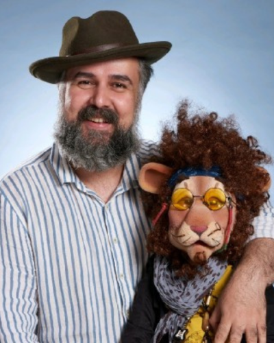 Portrait of Ali Etesamifar, a beared middle-eastern man wearing a hat with his arm around a lion puppet.