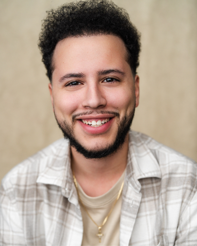 Portrait of Brandon Callado, a smiling hispanic college student, wearing a white and grey plaid collared shirt.