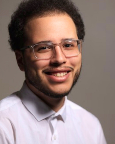 Portrait of Brandon Callado, a smiling hispanic college student, wearing wire-rimmed glasses and lavendar polo shirt.