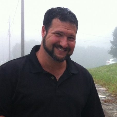 Porrait of a white man with dark hair and facial hair, wearning a dark polo shirt
