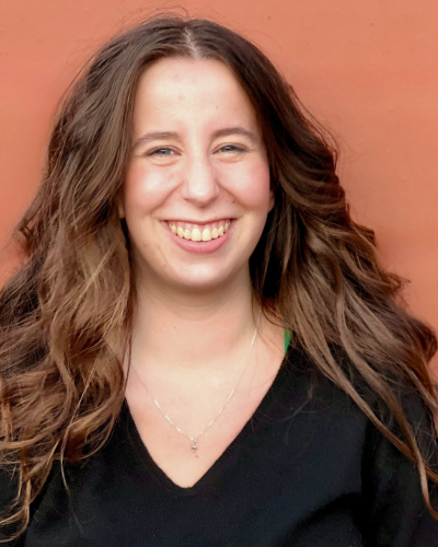 Portrait of Grace Darling, a white college student with long, think brown hair, smiling in front of an orange background.