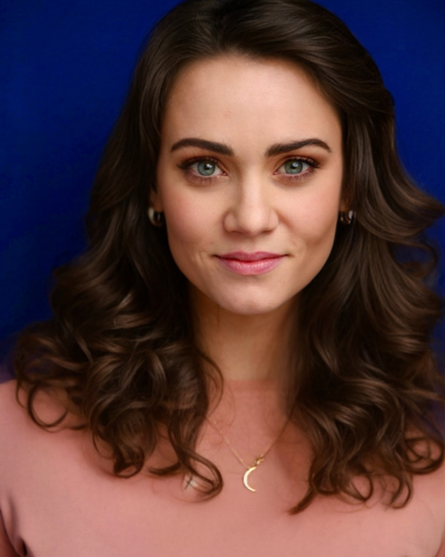 Portrait of Halli Gibson, a graduate student with long curly dark hair, wearning and pale rose colored top, standing in front of a royal blue backdrop..