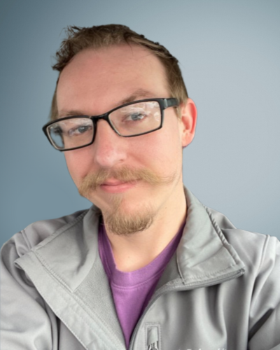 Portrait of Jake Neighbors, a white person with stylish rectangular glasses, blondish handlebar mustache and goatee.
