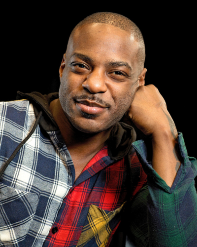 Portrait of a black graduate student with very short dark hair and a mustache, wearing a plaid shirt, with his cheek leaning on his fist.