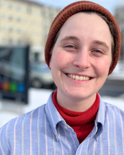 Portrait of Miranda Fyfe, a white graduate student with rosy cheeks, wearing a knit cap and blue/white striped button down shirt