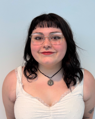 Portrait of a smiling white student with medium length dark hair and bangs, wearing wired-rimmed glasses and a white tank top.