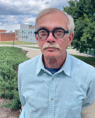 Portrait of an older gentleman with thick rimmed round glasses and white mustache, wearing a blue button down shirt.