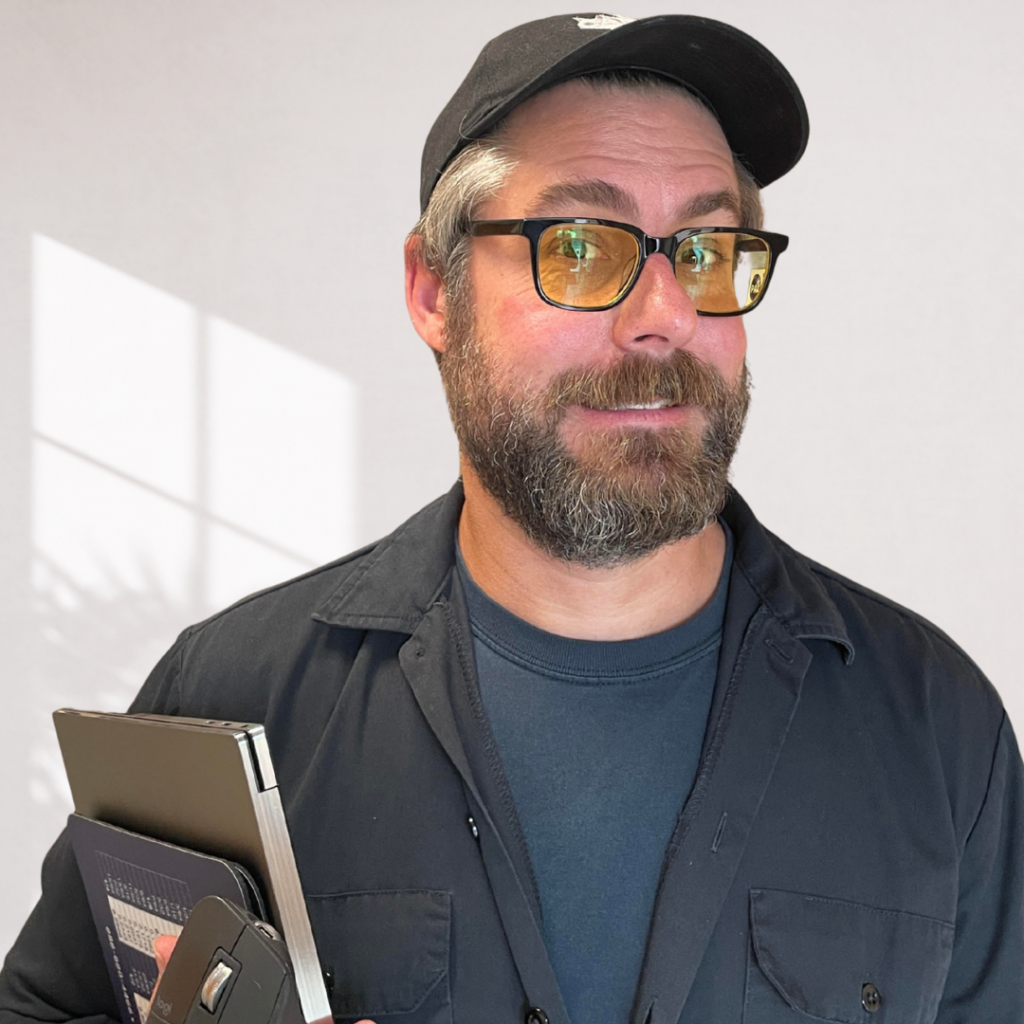 Portrait of a man with full beard & dark glasses, wearing a black baseball cap.