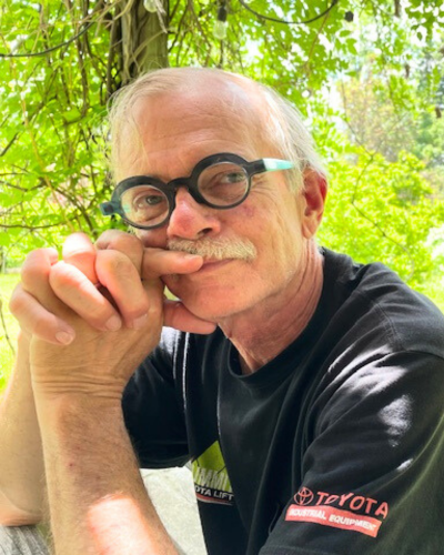 Portrait of an older gentleman with thick rimmed round glasses and white mustache, wearing a black t-shirt