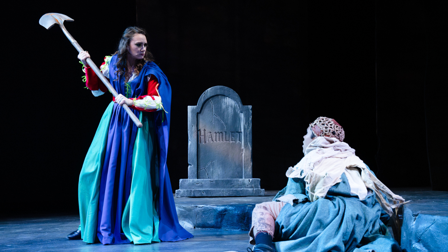 Ophelia, standing in from of Hamlet's tombstone, wields a weapon overhead as a zombie approaches.