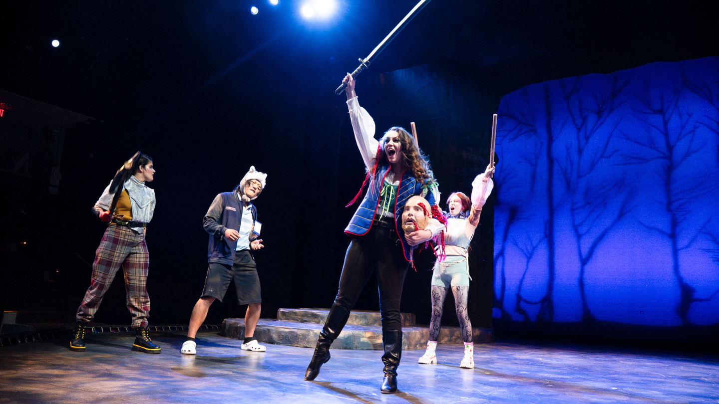 Lady M, Juliet, and Caliban look on as a victorious Ophelia carries the severed head of Fortenbras.