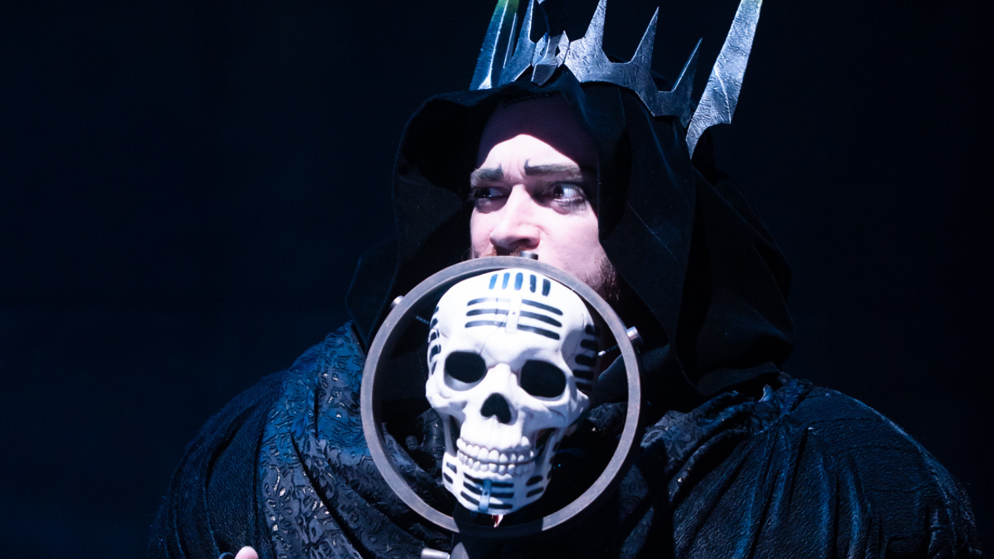 A ghost, wearing a large metal crown, speaks into a microphone carved from a scull.