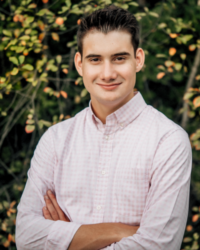 Portrait of a white student with combed back dark hair, wearing a light button-down shirt, smiling at the camera.