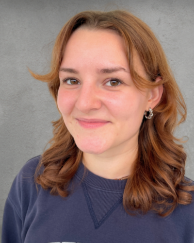 Portrait of Emily Hopkins, an undergraduate student with long wavy auburn hair, wearing a blue t-shirt, and smiling at hte camera.