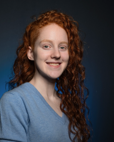 Portrait of an white undergraduate student with long red, tight curls, wearing a blue sweater.