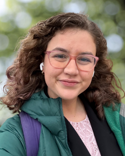 Portrait of an undergraduate student with medium legnth curly hair and pink glasses, wearing a green puffer coat.