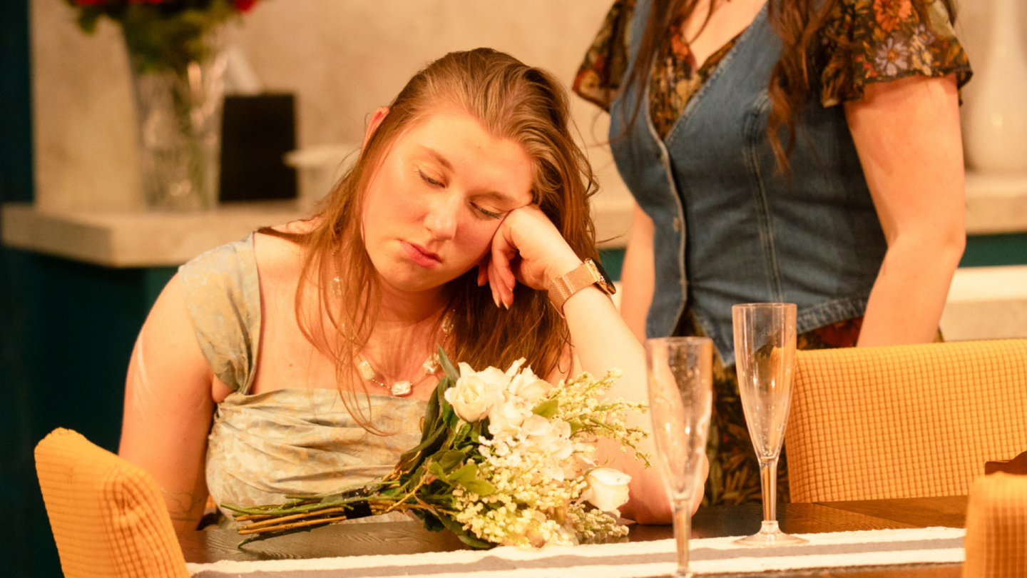Polycule Archive 9 In a scene from The Polycule, a young woman sits slumped over at a kitchen table, looking depressed while wearing a fancy green tafetta dress.