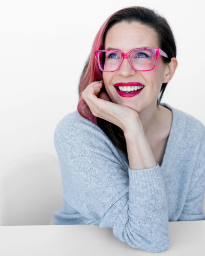 Portrait of a white woman with straight brown hair with a pink streak, wearing a light blue sweater and bright pink glasses, smiling and leaning her chin on her right hand.
