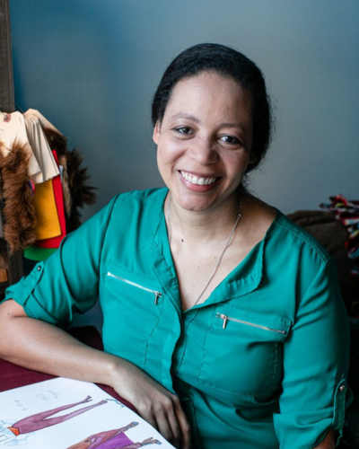 Portrait of a Black woman wearing a dark teal blouse, seated at a table, smiling at the camera.