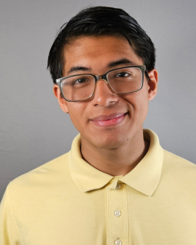 A person wearing glasses and a yellow polo shirt smiles against a gray background.