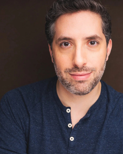A close-up portrait of a person with dark hair and a beard, wearing a dark blue henley shirt against a brown background.