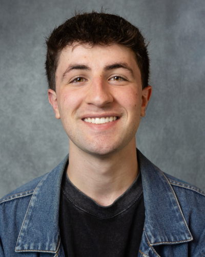 A young person with dark curly hair smiles broadly, wearing a denim jacket over a black t-shirt against a gray textured background.