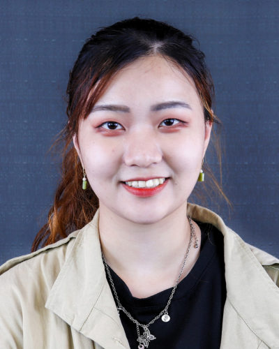 A woman with long brown hair pulled back smiles at the camera, wearing a light-colored jacket over a black shirt and a silver necklace.