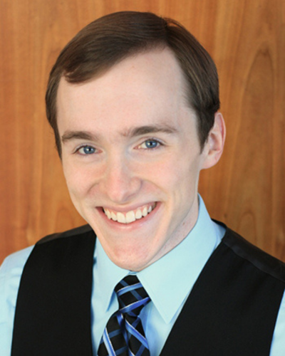 A man with brown hair and blue eyes smiles broadly, wearing a light blue collared shirt, a black vest, and a blue and black striped tie. The background is a warm, wooden surface with a wood grain texture.