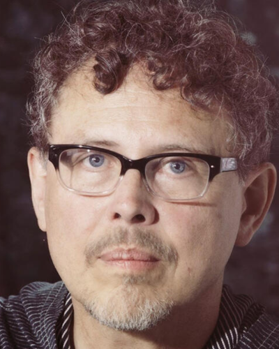 Close-up portrait of a person wearing black-rimmed glasses with curly hair and a mustache.