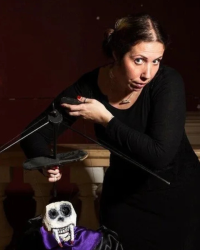 A performer in a dark outfit crouches onstage, holding a marionette of a small dog with a purple vest sitting in front, lit by warm stage lighting.