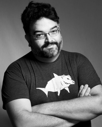 a black and white portrait of a person wearing glasses and a t-shirt with a shark-pig hybrid logo, arms crossed.