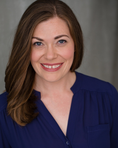 a headshot of a woman with brown hair, blue eyes, and a bright smile, wearing a dark blue blouse against a gray background.
