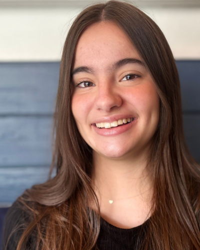 Smiling portrait of a person with long brown hair, wearing a dark top, outdoors in front of a blue-gray wall.